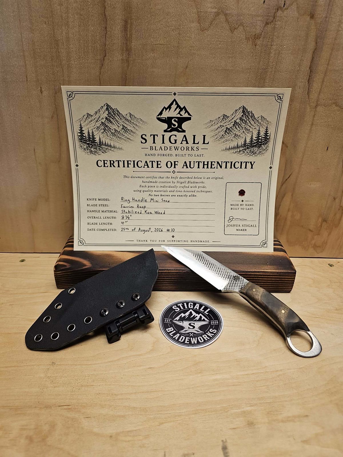 Ring-handle mini seax with a rasp-textured blade and koa handle beside its Kydex sheath and a Stigall Bladeworks sticker, in front of its signed certificate of authenticity